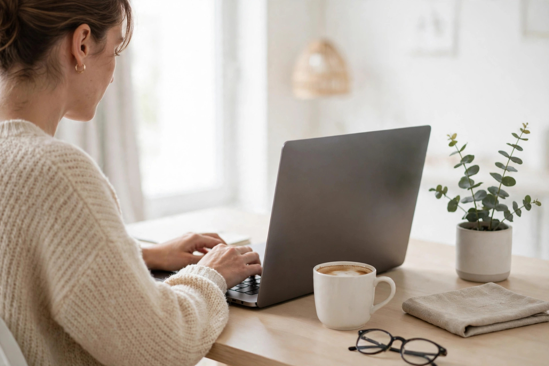 A woman typing on a laptop with a coffee at a bright home office desk.