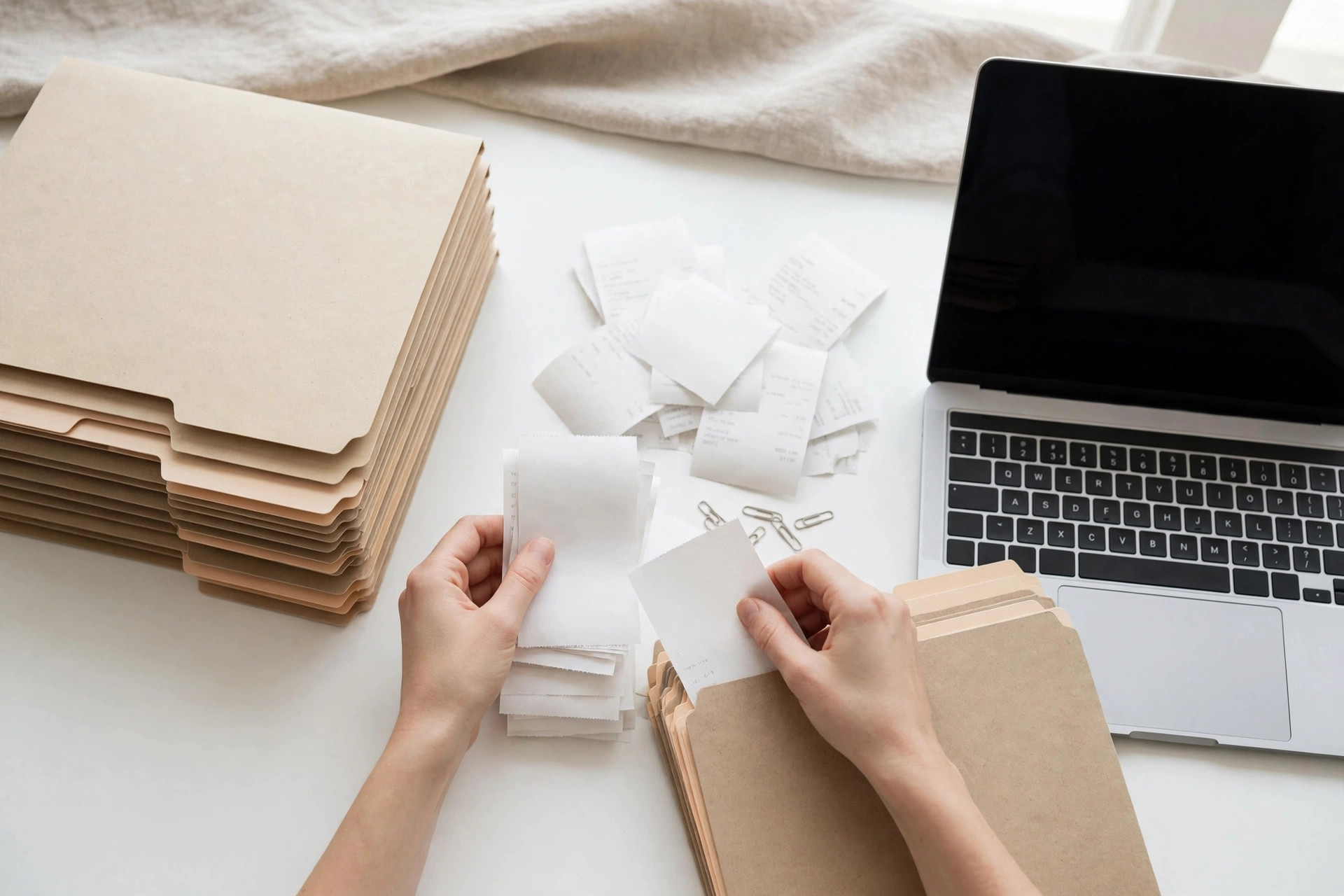 Hands organizing file folders and receipts beside a laptop